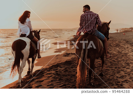 A loving couple in summer clothes riding a horse on a sandy beach at sunset. Sea and sunset in the background. Selective focus A loving couple in summer clothes riding a horse on a sandy beach at sunset. Sea and sunset in the background. Selective focus 95464288