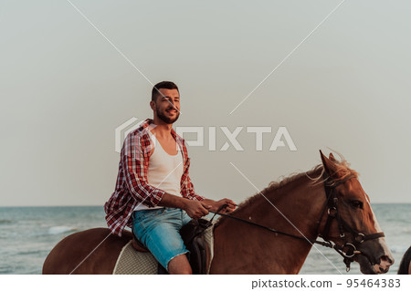 A loving couple in summer clothes riding a horse on a sandy beach at sunset. Sea and sunset in the background. Selective focus  95464383