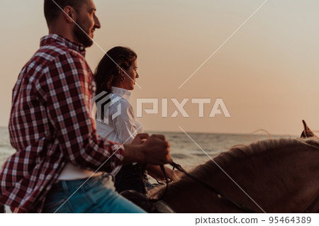A loving couple in summer clothes riding a horse on a sandy beach at sunset. Sea and sunset in the background. Selective focus A loving couple in summer clothes riding a horse on a sandy beach at sunset. Sea and sunset in the background. Selective focus 95464389