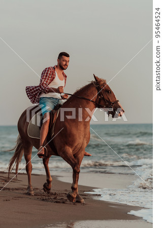 A modern man in summer clothes enjoys riding a horse on a beautiful sandy beach at sunset. Selective focus  95464524