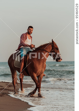 A modern man in summer clothes enjoys riding a horse on a beautiful sandy beach at sunset. Selective focus A modern man in summer clothes enjoys riding a horse on a beautiful sandy beach at sunset. Selective focus 95464529