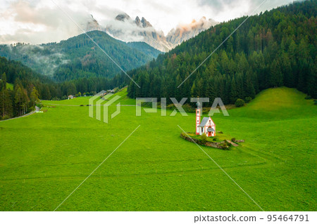 Aerial view of the Church of St. Johann against the Geisler peaks covered with clouds in Santa Maddalena village, Val Di Funes, Italy. Aerial view of the Church of St. Johann against the Geisler peaks covered with clouds in Santa Maddalena village, Val Di Funes, Italy. 95464791