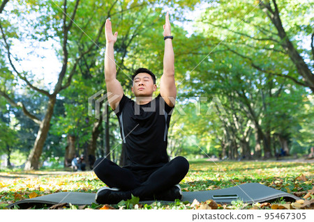 Asian man in the park on a sports mat sits in the lotus position, sportsman performs breathing exercises on a sunny day, man in a sports suit rests after physical and fitness exercises 95467305