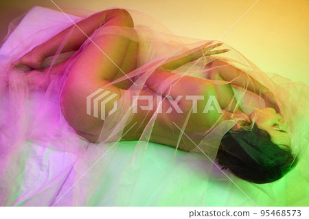 Portrait of tender woman covered in transparent fabric isolated over orange background in green pink neon light Portrait of tender woman covered in transparent fabric isolated over orange background in green pink neon light 95468573