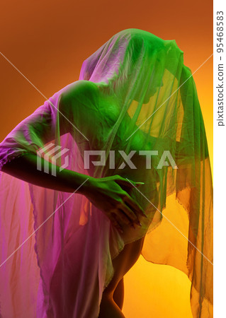 Silhouette of tender woman covered in transparent fabric isolated over orange background in green neon light 95468583