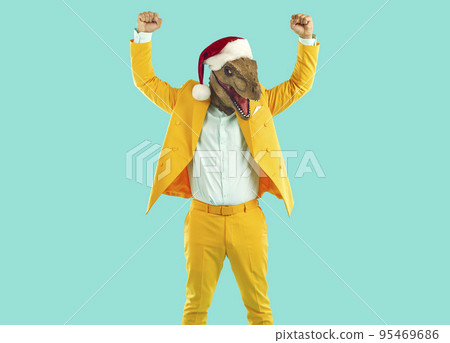 Funny man in yellow suit, dinosaur mask and Christmas hat raising fists up like a winner 95469686