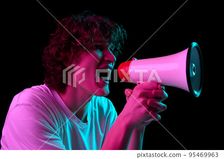 Closeup portrait of excited young man shouting at megaphone isolated over dark background in neon. Sport, betting, sales, news 95469963
