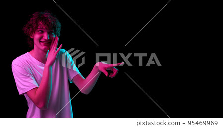 Portrait of young smiling man whispering isolated over dark background in purple neon light. Mental health, news, emotions, youth, sales, ad 95469969