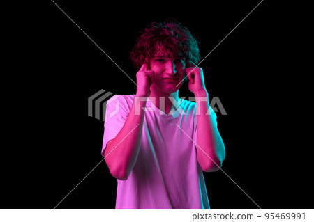 Terrible headache. Young man with discontent and undercurrent facial expression covering his ears isolated over dark background in neon light. Terrible headache. Young man with discontent and undercurrent facial expression covering his ears isolated over dark background in neon light. 95469991