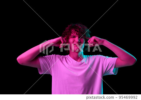 Looks annoyed. Young man with discontent and undercurrent facial expression covering his ears isolated over dark background in neon light. Looks annoyed. Young man with discontent and undercurrent facial expression covering his ears isolated over dark background in neon light. 95469992