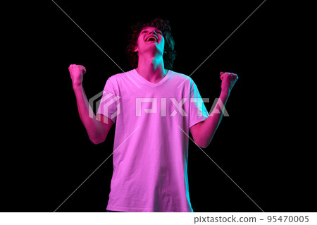 Half-length portrait of young man with winner emotions hands up isolated over dark background in pink neon light. Emotions, youth, sales, ad 95470005
