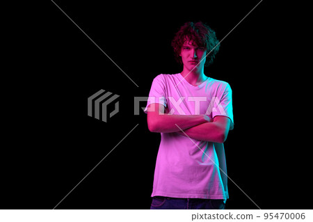 Angry young man standing with crossed arms isolated over dark background in purple neon light. Emotions, youth, sales, ad Angry young man standing with crossed arms isolated over dark background in purple neon light. Emotions, youth, sales, ad 95470006