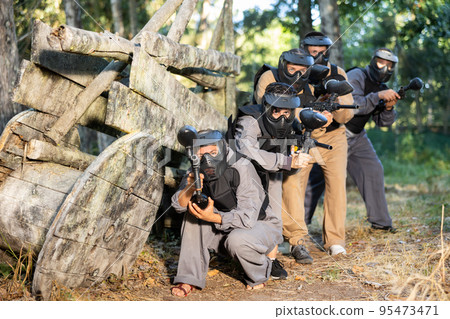 Group of paintball players aiming and shooting with guns outdoors Group of paintball players aiming and shooting with guns outdoors 95473471