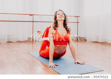 Middle aged well looking woman fitness instructor in Santas hat and red Sportswear stretching in the gym before her pilates, on a yoga mat near the large window on a sunny day, fitness routine concept 95474322