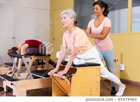 Hispanic woman pilates instructor helping aged woman exercising on wunda chair 95474460