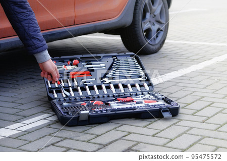 Mechanical tools box. Car driver using different repair tools for repairing and service an orange car. Tool set near the auto. Closeup. Mechanical tools box. Car driver using different repair tools for repairing and service an orange car. Tool set near the auto. Closeup. 95475572