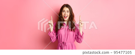 Beauty and fashion. Happy woman looking with amazement at copy space on top, pointing fingers up, smiling amazed, checking out promo offer, standing over pink background 95477105