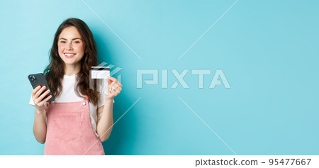 Young smiling woman making online payment, shop online with credit card and smartphone in hands, looking satisfied at camera, standing over blue background Young smiling woman making online payment, shop online with credit card and smartphone in hands, looking satisfied at camera, standing over blue background 95477667