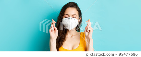 Health, covid-19 and lockdown concept. Close up of hopeful young woman in respirator mask, cross fingers and making wish, praying with eyes closed, standing against blue background Health, covid-19 and lockdown concept. Close up of hopeful young woman in respirator mask, cross fingers and making wish, praying with eyes closed, standing against blue background 95477680
