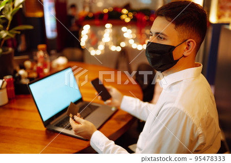 Young man wearing medical face mask working on laptop computer and speaks on the phone. Young man wearing medical face mask working on laptop computer and speaks on the phone. 95478333