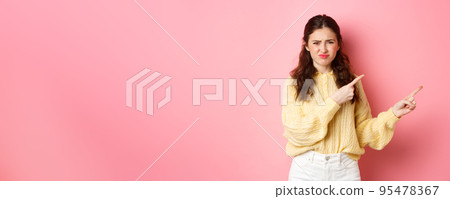 Skeptical and displeased young woman frowning, grimacing and pointing fingers right at copyspace, express dislike and disappointment, standing against pink background Skeptical and displeased young woman frowning, grimacing and pointing fingers right at copyspace, express dislike and disappointment, standing against pink background 95478367