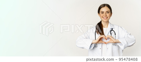 Portrait of smiling doctor, female physician showing heart sign and looking with care, love her patients, standing over white background Portrait of smiling doctor, female physician showing heart sign and looking with care, love her patients, standing over white background 95478408