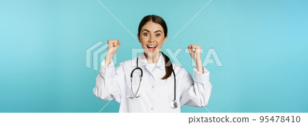 Enthusiastic doctor woman, rejoicing, winning and celebrating, achieve goal, standing in white coat against blue background Enthusiastic doctor woman, rejoicing, winning and celebrating, achieve goal, standing in white coat against blue background 95478410