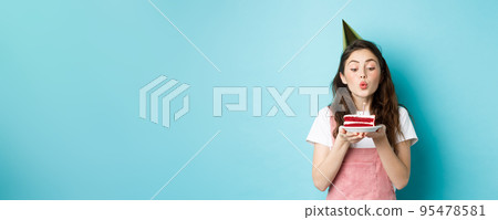 Holidays and celebration. Excited woman celebrating birthday, blowing candle on cake, wearing party cake and having fun, standing over blue background Holidays and celebration. Excited woman celebrating birthday, blowing candle on cake, wearing party cake and having fun, standing over blue background 95478581