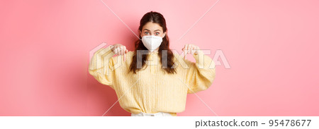 Preventive measures, health care concept. Excited woman in medical respirator pointing fingers down at copyspace, showing promo, standing against pink background Preventive measures, health care concept. Excited woman in medical respirator pointing fingers down at copyspace, showing promo, standing against pink background 95478677