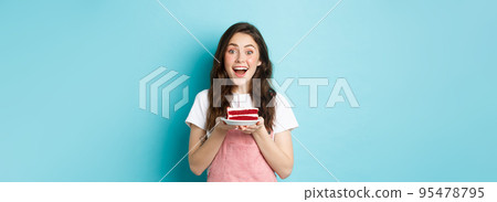 Holidays and party concept. Excited cute girl blowing candle on birthday cake, celebrating her b-day, standing over blue background Holidays and party concept. Excited cute girl blowing candle on birthday cake, celebrating her b-day, standing over blue background 95478795