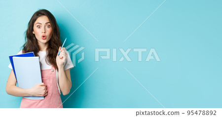 Young female student got an idea, raising hand with pen and look excited at camera, suggest plan, holding notebooks with study material, standing over blue background 95478892
