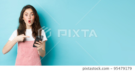 Portrait of excited woman pointing finger at smartphone, telling about online promo offer, showing advertisement on phone, standing against blue background 95479179