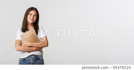 Portrait of smiling nostalgic female student looking upper left corner, holding notebooks Portrait of smiling nostalgic female student looking upper left corner, holding notebooks 95479224