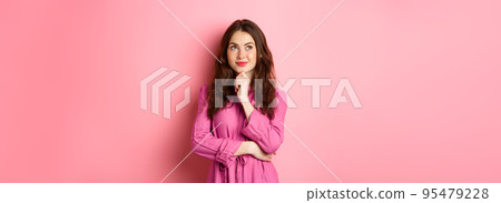 Stylish thoughtful woman with make up and spring dress, looking aside at logo and smiling pleased, thinking while standing against pink background 95479228