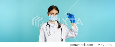Vaccination from covid and healthcare concept. Young woman doctor, nurse in face mask and gloves, using syringe to do vaccine shot, standing over torquoise background 95479229