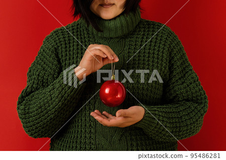 Asian woman in a green sweater on a red background holds a red Christmas ball in her hands 95486281