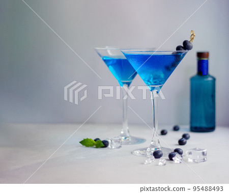 Blue cocktails in martini glass with ice cubes, mint and bluberries and bottle of gin 95488493