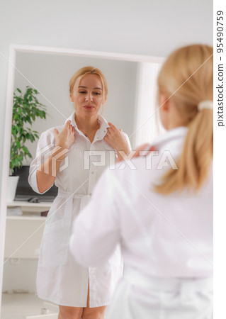 A blonde woman in white formal clothes looks at herself in the mirror. On a white background, green leaves at the back. A blonde woman in white formal clothes looks at herself in the mirror. On a white background, green leaves at the back. 95490759