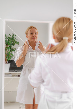 A blonde woman in white formal clothes looks at herself in the mirror. On a white background, green leaves at the back. A blonde woman in white formal clothes looks at herself in the mirror. On a white background, green leaves at the back. 95490760