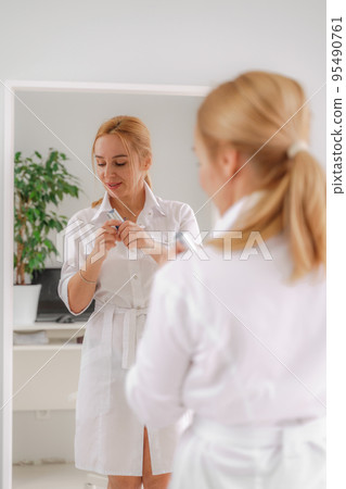 A blonde woman in white formal clothes looks at herself in the mirror. On a white background, green leaves at the back. A blonde woman in white formal clothes looks at herself in the mirror. On a white background, green leaves at the back. 95490761