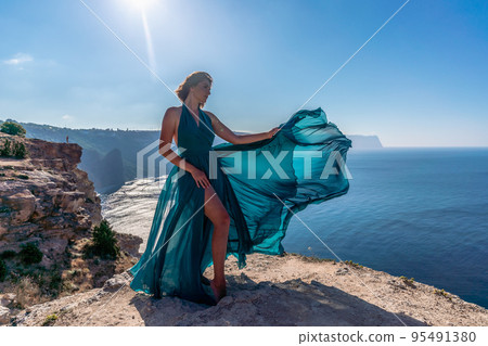 A girl with loose hair in a long mint dress descends the stairs between the yellow rocks overlooking the sea. A rock can be seen in the sea. Sunny path on the sea from the rising sun A girl with loose hair in a long mint dress descends the stairs between the yellow rocks overlooking the sea. A rock can be seen in the sea. Sunny path on the sea from the rising sun 95491380