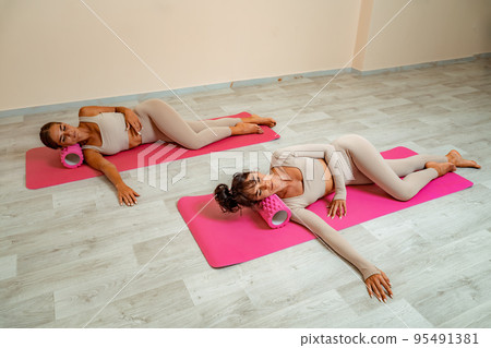 Fit athletic women in beige sportswear ride on a pink foam myofascial massage roller, massaging their muscles on the floor of a yoga mat in the gym. Active girls do a fitness workout in the gym. MFR Fit athletic women in beige sportswear ride on a pink foam myofascial massage roller, massaging their muscles on the floor of a yoga mat in the gym. Active girls do a fitness workout in the gym. MFR 95491381