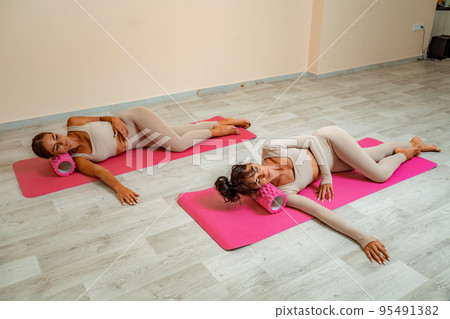 Fit athletic women in beige sportswear ride on a pink foam myofascial massage roller, massaging their muscles on the floor of a yoga mat in the gym. Active girls do a fitness workout in the gym. MFR Fit athletic women in beige sportswear ride on a pink foam myofascial massage roller, massaging their muscles on the floor of a yoga mat in the gym. Active girls do a fitness workout in the gym. MFR 95491382