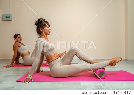 Fit athletic women in beige sportswear ride on a pink foam myofascial massage roller, massaging their muscles on the floor of a yoga mat in the gym. Active girls do a fitness workout in the gym. MFR Fit athletic women in beige sportswear ride on a pink foam myofascial massage roller, massaging their muscles on the floor of a yoga mat in the gym. Active girls do a fitness workout in the gym. MFR 95491417