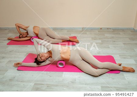 Fit athletic women in beige sportswear ride on a pink foam myofascial massage roller, massaging their muscles on the floor of a yoga mat in the gym. Active girls do a fitness workout in the gym. MFR Fit athletic women in beige sportswear ride on a pink foam myofascial massage roller, massaging their muscles on the floor of a yoga mat in the gym. Active girls do a fitness workout in the gym. MFR 95491427