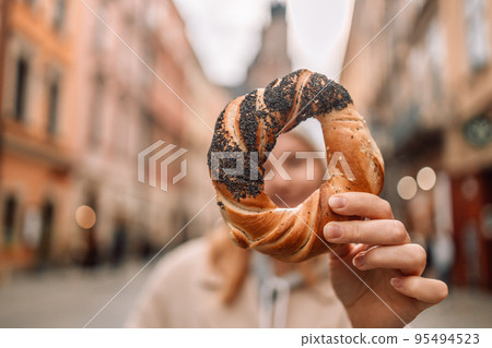 Happy young blonde woman tourist in stylish clothes holding pretzel obwarzanek on the market square in Krakow in Poland 95494523