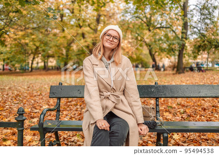 Autumn woman in a stylish beautiful warm coat from a knitted hat relaxes on a bench on a warm day 95494538