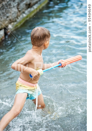 Small boy holds bright water-jet gun standing in sea Small boy holds bright water-jet gun standing in sea 95494649