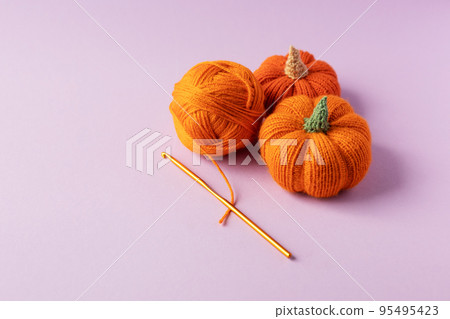 Knitted orange pumpkins and an orange ball of thread with a crochet hook on a purple background, autumn composition. Halloween concept. Knitted orange pumpkins and an orange ball of thread with a crochet hook on a purple background, autumn composition. Halloween concept. 95495423