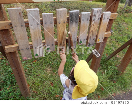 A child wearing a yellow hat is hitting outdoor wooden playground equipment with a wooden stick. 95497550